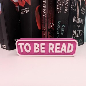 May include: A pink and white rectangular sign with the text "TO BE READ". The sign is in front of a row of books with black covers. The sign is a decorative item for book lovers, perfect for a bookshelf or desk.