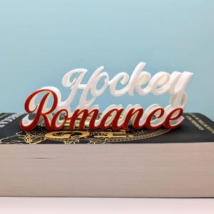 May include: A white and red "Hockey Romance" sign, in a cursive font, rests on a book with gold lettering. The sign is made of a 3D printed material. The background is a solid light blue.