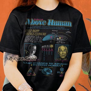 Puede incluir: Camiseta negra con un estampado gráfico de estilo retro que presenta el texto "Above Human" y un diagrama del sistema solar. El diseño también incluye un gráfico de un extraterrestre gris y un hombre con una expresión de sorpresa. El texto "Last Chance to Advance Beyond Human" también está incluido en el diseño.