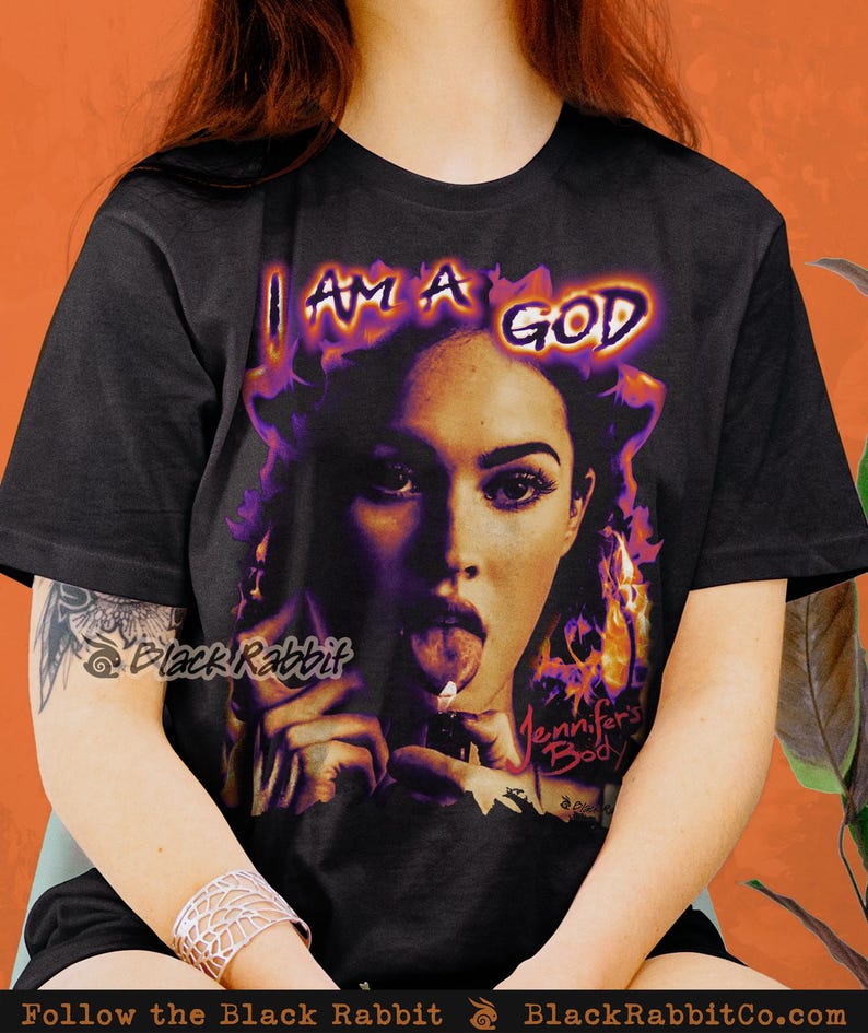 May include: Black t-shirt with a graphic print of a woman with a cigarette in her mouth. The text "I AM A GOD" is printed above her head and "Jennifer's Body" is printed below her. The graphic is in shades of purple and orange.