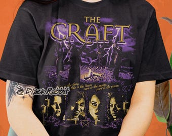 Könnte beinhalten: Schwarzes T-Shirt mit einem Grafikdruck des Films "The Craft". Die Grafik zeigt die vier Hauptfiguren des Films, mit dem Titel des Films in goldenen Buchstaben darüber. Der Text "the time, this is the hour, ours is the magic, curs is the power." ist unter der Grafik gedruckt.
