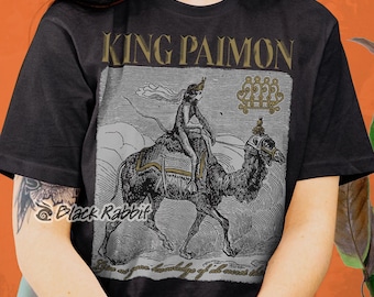May include: Black t-shirt with a gold print of a demon riding a camel. The print includes the text "King Paimon" and a sigil. The text "Give you knowledge of all secret things" is printed below the image.