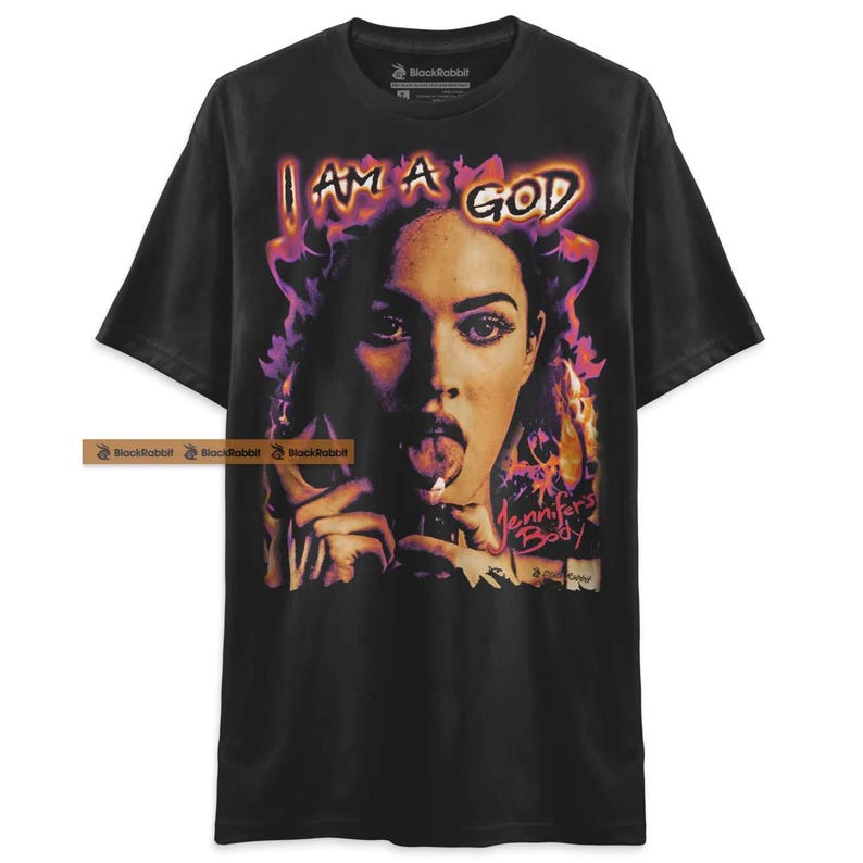 May include: Black t-shirt with a graphic print of a woman with a tongue sticking out and a cigarette in her hand. The text "I AM A GOD" is printed above the woman's head. The text "Jennifer's Body" is printed below the woman's head.