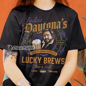 Puede incluir: Camiseta negra con un diseño gráfico de un bar llamado "Jackie Daytona's Lucky Brews Bar & Grill". El gráfico incluye una caricatura de un hombre sosteniendo una jarra de cerveza y el texto "Est. 2020", "Regular Human Bartender", "Go Bucks!" y "Clairton, Penn. Transylvania".