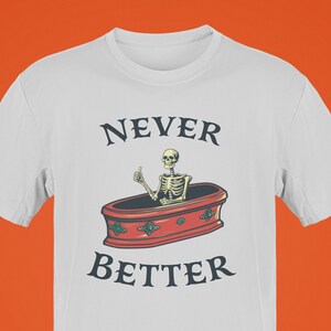 K&ouml;nnte beinhalten: Wei&szlig;es T-Shirt mit dem Text "NEVER BETTER" &uuml;ber einer Illustration eines Skeletts, das in einem Sarg einen Daumen hoch h&auml;lt. Der Sarg ist rot mit dekorativen Akzenten.