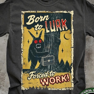 May include: Dark gray t-shirt with a vintage-style graphic. The graphic features a black monster with red eyes holding a briefcase, with the text "Born to LURK" and "Forced to WORK!" in a distressed font. A circular logo is at the bottom.