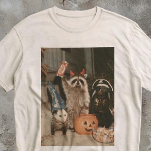 May include: A cream-coloured t-shirt featuring a graphic print of three animals dressed for Halloween: an opossum in a blue star-patterned hat, a raccoon with red devil horns holding a KitKat bar, and a skunk wearing a pirate hat and eye patch.