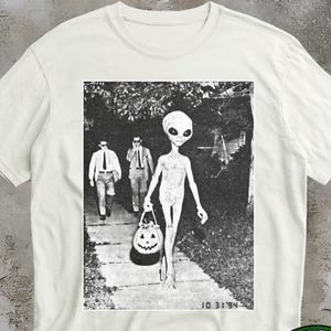 May include: A white t-shirt featuring a black and white graphic print of an alien walking on a pavement with two figures in suits behind it. The alien carries a jack-o'-lantern bucket. The text 10 31 94 is printed in the bottom right corner.