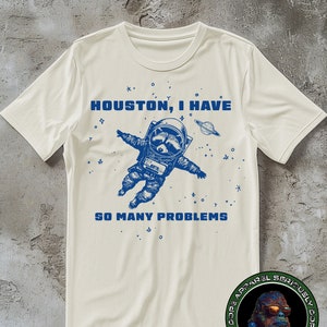 May include: A white t-shirt with a blue graphic of a raccoon in a spacesuit with the text "Houston, I have so many problems". The graphic is surrounded by stars and planets. The t-shirt has a green and black logo on the bottom right corner.