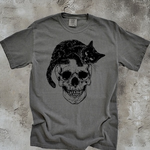 Puede incluir: Una camiseta gris con un gato negro sentado sobre un gr&aacute;fico de calavera. El gato mira hacia la derecha y la calavera tiene un dise&ntilde;o detallado.