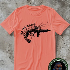 May include: A coral pink t-shirt featuring a black graphic of a shrimp holding a gun with the text "Bang Bang" printed on the front. The shirt has a round logo on the bottom right corner with the text "Dope Apparel Seriously Dope Apparel Seriously".