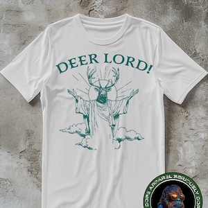 Peut inclure: T-shirt blanc avec une tête de cerf verte avec des bois et une auréole, avec le texte "DEER LORD!" au-dessus. Le cerf porte une robe et se tient sur un nuage. Le t-shirt a un logo en bas à droite qui dit "Dope Apparel Seriously Dope Apparel Seriously".