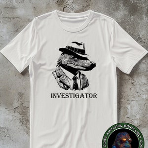 Könnte beinhalten: Weißes T-Shirt mit einer schwarz-weißen Illustration eines Alligators, der einen Fedora und eine Krawatte trägt. Der Text "INVESTIGATOR" ist unter der Illustration aufgedruckt.