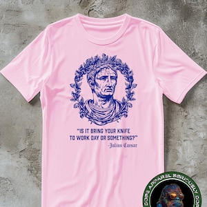 Puede incluir: Camiseta rosa con una imagen azul de Julio César con una corona de laurel y una cita: "IS IT BRING YOUR KNIFE TO WORK DAY OR SOMETHING?" -Julio César