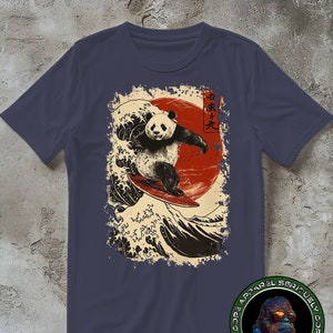 Puede incluir: Una camiseta azul marino con un gráfico de un oso panda surfeando en una ola con un sol rojo de fondo. El gráfico es de estilo japonés con caracteres kanji que se leen "波乗り天" (que significa "paraíso del surf").