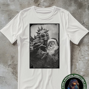 May include: A white t-shirt with a black and white image of a monster and Santa Claus taking a selfie. The image is in a vintage style frame. The bottom of the shirt has a black and white logo with a gorilla wearing sunglasses.