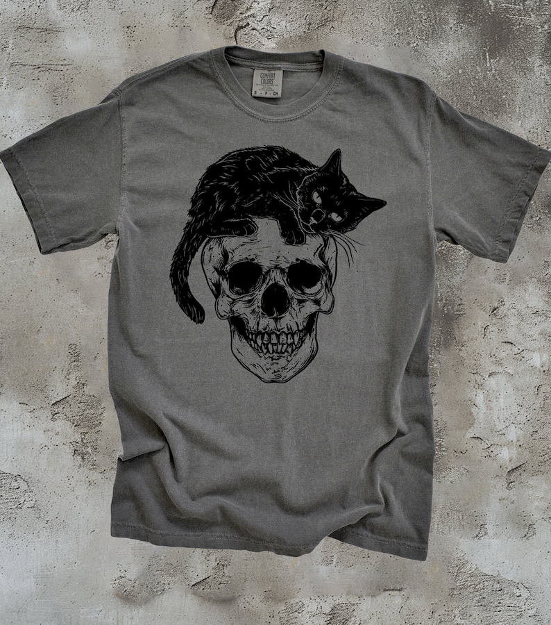 Puede incluir: Una camiseta gris con un gato negro sentado sobre un gr&aacute;fico de calavera. El gato mira hacia la derecha y la calavera tiene un dise&ntilde;o detallado.