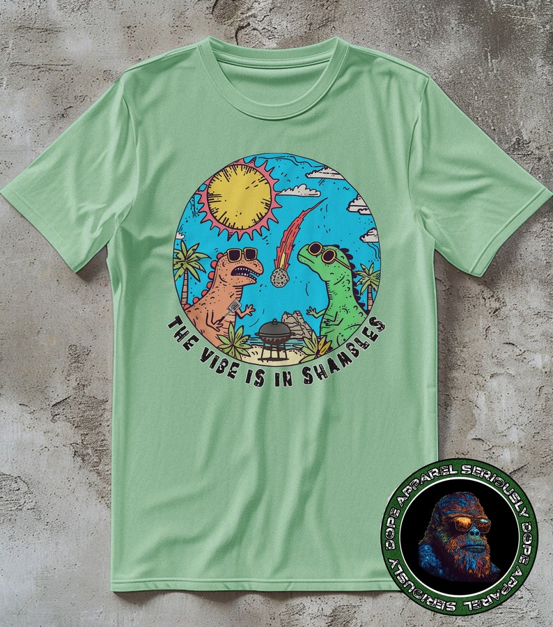 K&ouml;nnte beinhalten: Ein gr&uuml;nes T-Shirt mit einer kreisf&ouml;rmigen Grafik von zwei Dinosauriern, einer Sonne und einem Kometen. Der Text "The Vibe is in Shambles" ist um die Grafik herum geschrieben. Das Logo "Dope Apparel Seriously Dope Apparel" befindet sich in der rechten unteren Ecke.