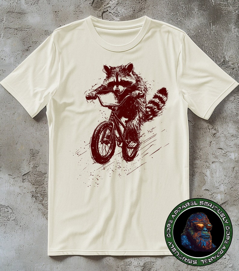 Pu&ograve; includere: Una t-shirt bianca con una grafica rossa di un procione che guida una bicicletta. La grafica &egrave; in stile vintage con un effetto grunge. La t-shirt ha un logo rotondo in basso a destra che dice "Dope Apparel Seriously Dope Apparel Seriously".