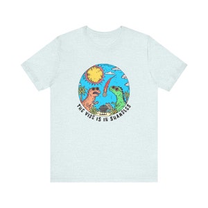 K&ouml;nnte beinhalten: Hellblaues T-Shirt mit einem Motiv von zwei Dinosauriern, die Sonnenbrillen tragen und grillen. Der Text "The vibe is in shambles" ist um das Motiv herum geschrieben.