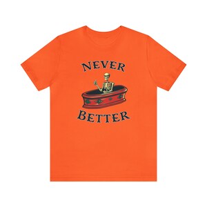 K&ouml;nnte beinhalten: Orangefarbenes T-Shirt mit dem schwarzen Text "NEVER BETTER" und einer Illustration eines Skeletts, das aus einem Sarg einen Daumen hoch zeigt. Das Design ist mittig platziert.