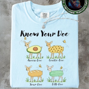Puede incluir: Camiseta Comfort Colors azul claro con la frase "Know Your Doe" e ilustraciones de ciervos con cuerpos temáticos de comida: aguacate, galleta, pan y eneldo. Se ve un collar plateado con un colgante.