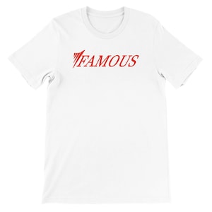 Puede incluir: Una camiseta blanca con la palabra "FAMOUS" impresa en rojo en el pecho. La palabra está en una fuente estilizada. La camiseta tiene mangas cortas y cuello redondo.