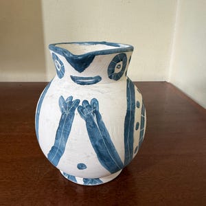 Könnte beinhalten: Ein weißer Keramikkrug mit handgemalten blauen Mustern. Die Außenseite zeigt stilisierte blaue Figuren mit erhobenen Armen, kreisförmige Muster am Rand und vertikale Streifen. Der Krug hat einen bauchigen Körper und einen breiten, offenen Ausguss.