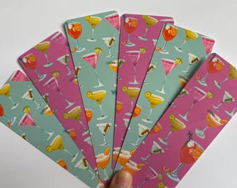 May include: A collection of cocktail-themed bookmarks. Each bookmark features a different cocktail design in shades of pink, yellow, orange, and white against a teal or pink background. The bookmarks are arranged in a fan shape.