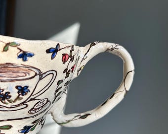May include: Cream-colored ceramic mug with a floral design. The mug features a hand-painted illustration of a teacup and saucer, with blue, red, and green floral accents. The handle is adorned with a delicate vine pattern.