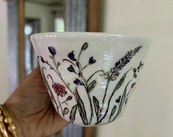 May include: A white ceramic bowl featuring a hand-painted botanical design with delicate black outlines. The artwork displays various wildflowers in shades of blue, red, and green, including tall stems, small buds, and leafy foliage scattered across the exterior surface.