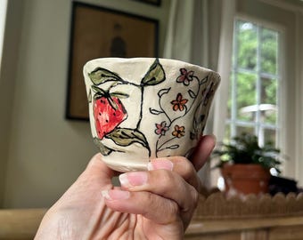 May include: A small, handmade ceramic cup with a cream-colored base. The cup is decorated with a vibrant red strawberry, green leaves, and delicate pink and orange flowers. The design is outlined in black.