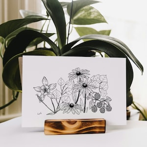 May include: Black and white line art print of a bouquet of flowers, including black-eyed susans, a single white flower, and blackberries. The print is displayed on a wooden easel.