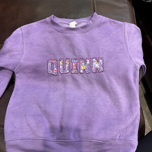 May include: A lavender crewneck sweatshirt featuring the name QUINN appliqued across the chest in block letters. The letters are filled with a colorful floral pattern consisting of small pink, yellow, blue, and white flowers on a light purple background.