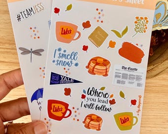 May include: Two white sticker sheets titled Gilmore Stickers Sheet featuring various illustrations including coffee mugs labeled Luke's, stacks of pancakes, a book, a newspaper, a Yale University pennant, and text reading I smell snow and Where you lead I will follow.