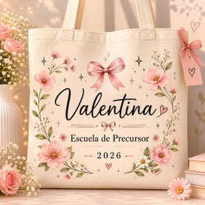 May include: A beige canvas tote bag featuring the name "Valentina" in elegant script, surrounded by pink floral illustrations and a pink bow. The text "Escuela de Precursor - 2026" is also displayed. The bag has sturdy handles and a decorative tag.