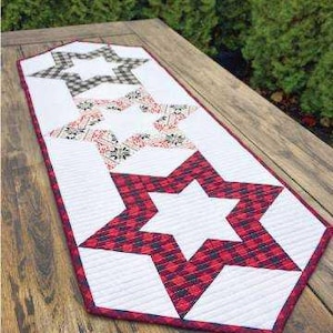 May include: A quilted table runner with a star pattern. The runner features white, red, and black plaid star designs. The edges are trimmed with a red border. The runner is placed on a wooden table.
