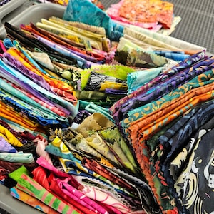 May include: A gray plastic bin filled with a large collection of folded fabric scraps in a wide variety of colors and patterns, including bright pinks, oranges, blues, greens, and purples, with some pieces featuring intricate batik-style designs.