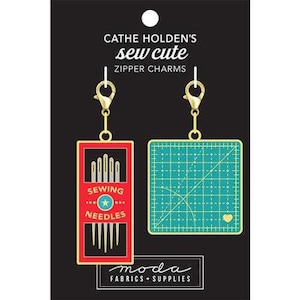 May include: Two sewing-themed zipper charms. One charm is a red rectangle with the words "SEWING NEEDLES" and a star, with needles inside. The other is a teal cutting mat with a gold heart. Both have gold-colored clasps.
