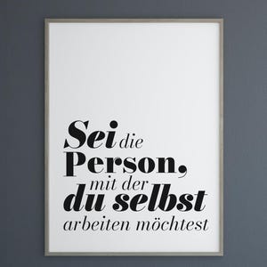 May include: A framed white poster with black text that reads "Sei die Person, mit der du selbst arbeiten möchtest." The poster is hanging on a dark gray wall. A digital download icon is in the upper left corner.