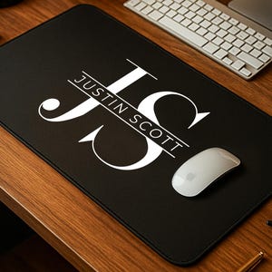 May include: A black rectangular desk mat with the name "Justin Scott" in white script. The mat features a large white "JS" monogram. A white computer mouse rests on the mat, with a keyboard and monitor in the background.