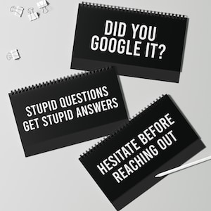 以下が含まれることがあります： 「DID YOU GOOGLE IT?」、「STUPID QUESTIONS GET STUPID ANSWERS」、および「HESITATE BEFORE REACHING OUT」という白文字の3つの黒い卓上カレンダー。いくつかのシルバーのペーパークリップが散らばっています。