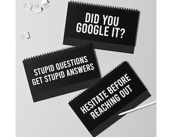 May include: Three black desk calendars with white text: "DID YOU GOOGLE IT?", "STUPID QUESTIONS GET STUPID ANSWERS", and "HESITATE BEFORE REACHING OUT". Several silver binder clips are scattered around.