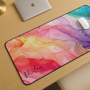 May include: A large rectangular mousepad with a colorful abstract design. The design features flowing patterns in shades of pink, orange, purple, and teal, with gold accents. The name "Miley" is written in cursive on the lower left corner. A white mouse sits on the pad.