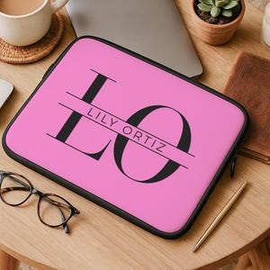 Könnte beinhalten: Eine pinkfarbene Laptoptasche mit dem Namen "LILY ORTIZ" in schwarzer Schrift. Die Hülle hat einen schwarzen Reißverschluss und eine schwarze Umrandung. Das Bild befindet sich auf einem Holztisch mit einer Kaffeetasse, einer Maus, einer Brille und anderen Gegenständen.