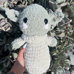 May include: A handmade, light green and cream-colored crocheted doll with large black eyes, large ears, and small pink cheeks. The doll is held in front of a background of green plants.
