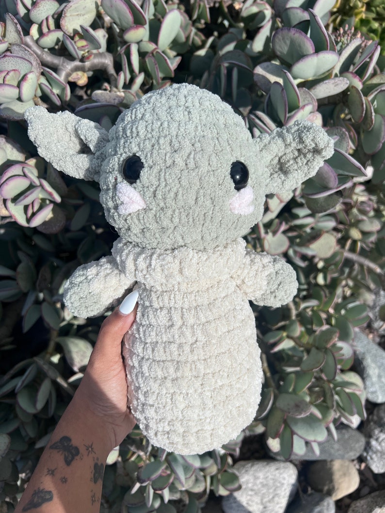 May include: A handmade, light green and cream-colored crocheted doll with large black eyes, large ears, and small pink cheeks. The doll is held in front of a background of green plants.