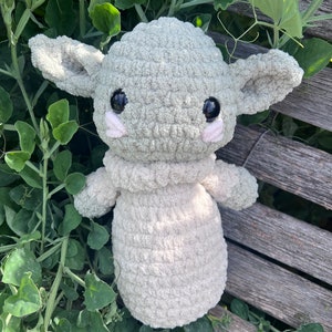 May include: A crocheted plush toy in the likeness of a character with large ears and big black eyes. The toy is primarily light green with a cream-colored body and small pink accents. It is resting on a wooden surface surrounded by green foliage.