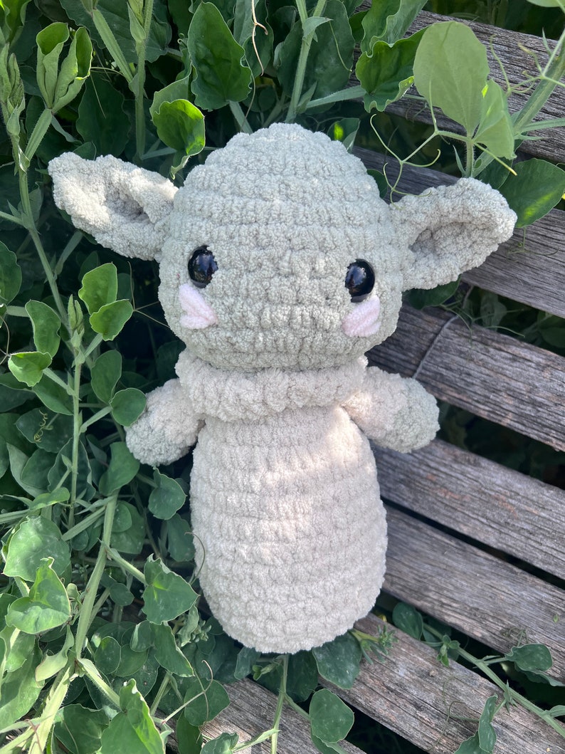 May include: A crocheted plush toy in the likeness of a character with large ears and big black eyes. The toy is primarily light green with a cream-colored body and small pink accents. It is resting on a wooden surface surrounded by green foliage.