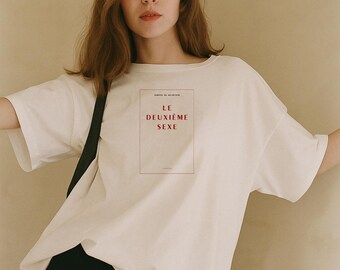 Op de afbeelding: Een off-white t-shirt met korte mouwen en een rechthoekige grafische print op de borst. De print bevat de tekst LE DEUXIÈME SEXE in rode letters, omsloten door een dunne rode rand, met kleine tekst erboven en eronder.
